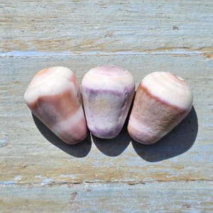 May include: Three polished stones with a smooth, rounded shape. The stones have a mix of colors, including white, purple, and brown, with a striped pattern. The stones are arranged in a row on a light blue and brown wooden surface.