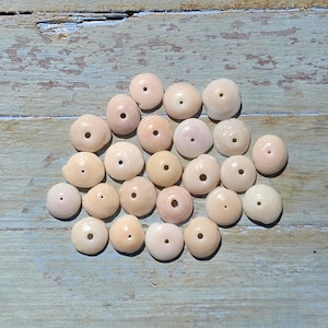 May include: A collection of light beige, round beads with a central hole, scattered on a weathered, light blue wooden surface. The beads vary slightly in color, with some having a pinkish hue. These beads are likely for jewelry making.