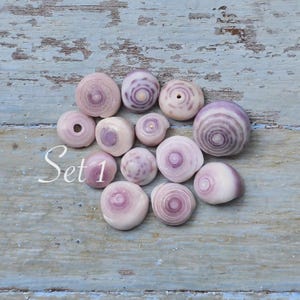 Peut inclure: Un ensemble de treize petits coquillages ronds et lisses, chacun présentant un motif en spirale distinct dans des tons de rose pâle, lavande et blanc. Les coquillages sont disposés en groupe, avec le texte Set 1 écrit en blanc.