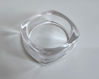 Puede incluir: Un brazalete transparente de forma cuadrada. El material acrílico transparente refleja la luz, creando reflejos y sombras. El brazalete tiene un acabado liso y pulido y un diseño moderno y minimalista. Este accesorio es adecuado para el uso diario.