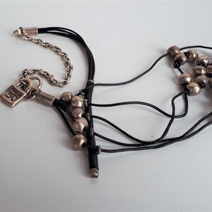 Pode incluir: Um fecho de cadeado prateado com a palavra "OG" nele pendurado em um cord&atilde;o de couro preto com contas prateadas. O cord&atilde;o est&aacute; amarrado a uma corrente prateada.
