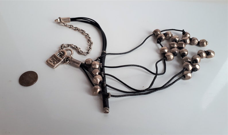 Pode incluir: Um fecho de cadeado prateado com a palavra "OG" nele pendurado em um cord&atilde;o de couro preto com contas prateadas. O cord&atilde;o est&aacute; amarrado a uma corrente prateada.