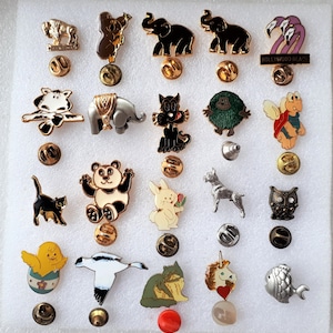 Pode incluir: Uma coleção de 20 pinos de esmalte com vários personagens de animais e desenhos animados. Os pinos são exibidos em um fundo branco. Alguns dos pinos incluem um panda, um gato, um coelho, um peixe, um pássaro, um unicórnio e um flamingo.