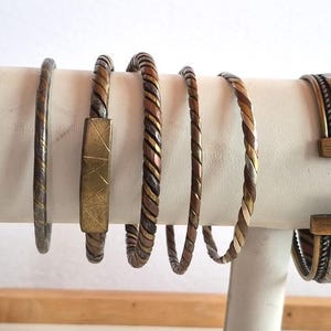 May include: A selection of mixed metal bracelets on a white stand. The bracelets feature various designs, including twisted and solid styles. The colours include silver, copper, and brass. The stand is white.