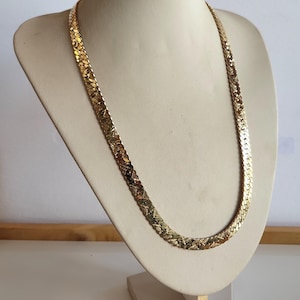 May include: A gold-coloured herringbone chain necklace displayed on a cream-coloured bust. The necklace has a flat, woven design. The bust is on a white surface.