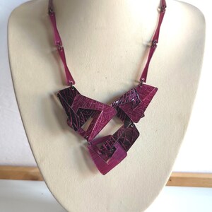 May include: A magenta-colored necklace with geometric shapes. The necklace is displayed on a mannequin. The necklace has a series of rectangular and square shapes connected by smaller, rectangular links. The shapes have a textured pattern.