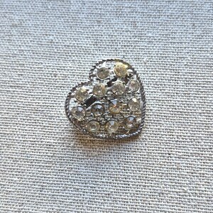 May include: A heart-shaped brooch with a silver-coloured metal frame. The brooch is encrusted with numerous clear, round rhinestones. The heart shape is outlined with a twisted rope design.