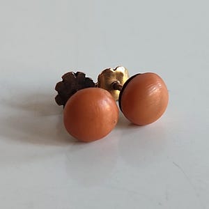 May include: A pair of round, peach-colored stud earrings. The earrings have a smooth, polished surface and are set on gold-toned posts with decorative backs. The earrings are displayed on a white surface.