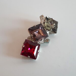 Peut inclure: Une broche en argent avec trois strass de forme carrée. Le strass du haut est vert, celui du milieu est violet et celui du bas est rouge.