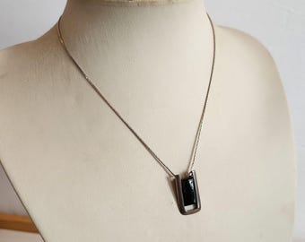 Può includere: Una collana color argento con un ciondolo rettangolare. Il ciondolo presenta un inserto rettangolare scuro, forse nero, all'interno di una cornice argentata. La collana è esposta su un manichino.