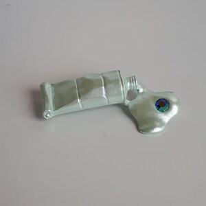 May include: A silver paint tube with a green and blue iridescent gem, squeezed out onto a white surface.