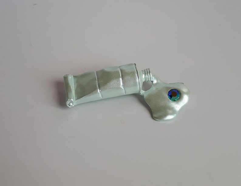 May include: A silver paint tube with a green and blue iridescent gem, squeezed out onto a white surface.
