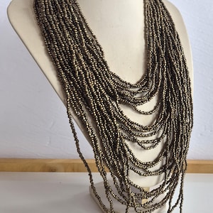 May include: A multi-strand necklace made of small, bronze-coloured beads. The necklace is displayed on a mannequin bust, highlighting its cascading layers and intricate design. The beads have a metallic sheen.