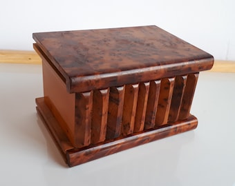 May include: A wooden puzzle box with a rectangular shape. The box is made of dark brown wood with a patterned top and vertical carved details on the sides. The box sits on a base and has a lid.