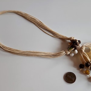 May include: A natural-toned necklace featuring a large, iridescent seashell pendant. The necklace has multiple strands of beige cord and is adorned with brown, green, and white beads. A penny is next to the necklace for size reference.