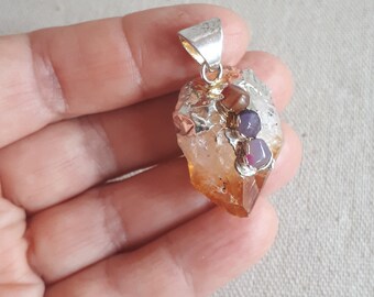 May include: A silver pendant with a large, rough-cut, orange-brown crystal. The crystal is accented with smaller, colorful stones in shades of red, purple, and brown.