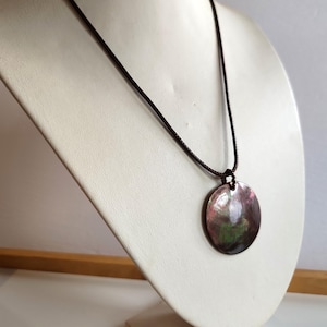 May include: A pendant necklace with a large, iridescent, round shell pendant. The pendant has pink, green, and purple hues. The necklace has a dark brown cord. The necklace is displayed on a white bust.