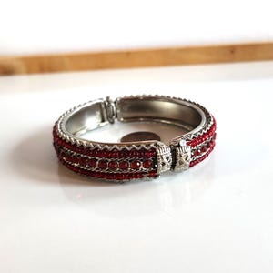 Puede incluir: Un brazalete de plata con detalles de cuentas rojas. El brazalete tiene un cierre de bisagra.