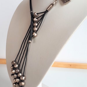 Pode incluir: Um colar longo de contas prateadas com um cord&atilde;o preto e um fecho de cadeado prateado. O colar &eacute; apresentado em um busto de manequim branco.