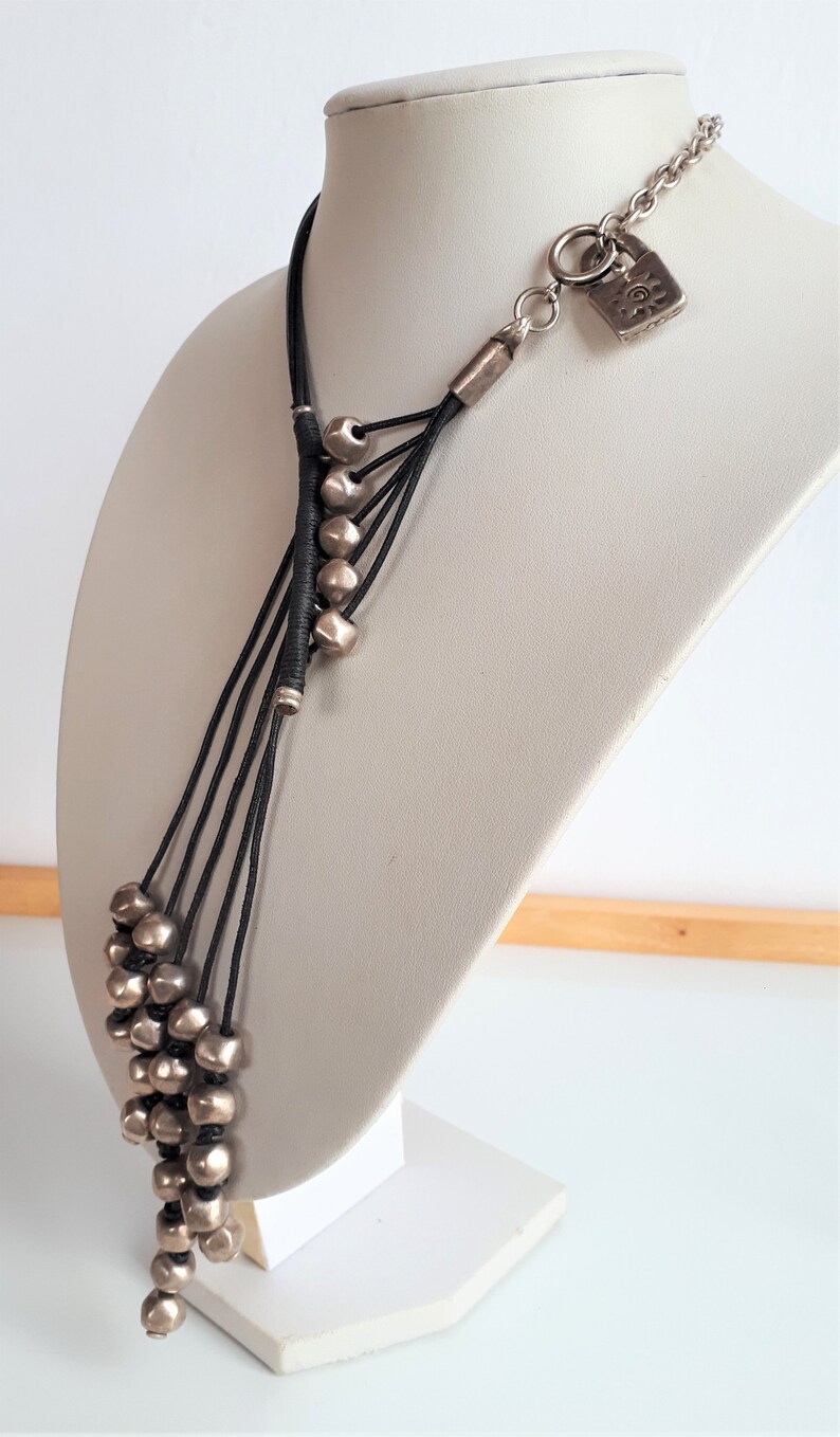 Pode incluir: Um colar longo de contas prateadas com um cord&atilde;o preto e um fecho de cadeado prateado. O colar &eacute; apresentado em um busto de manequim branco.