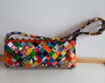 May include: A rectangular, woven handbag made from colorful recycled materials. The bag features a zipper closure with an orange lining and a matching woven handle. The bag's design incorporates various colors and patterns.