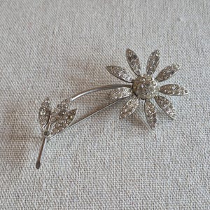 Könnte beinhalten: Silberfarbene Gänseblümchen-Brosche mit funkelnden, klaren Strasssteinen. Die Brosche zeigt eine detaillierte Blume mit mehreren Blütenblättern und einem gebogenen Stiel mit Blättern, die alle mit den gleichen schimmernden Steinen besetzt sind. Ein klassisches Blumenaccessoire.