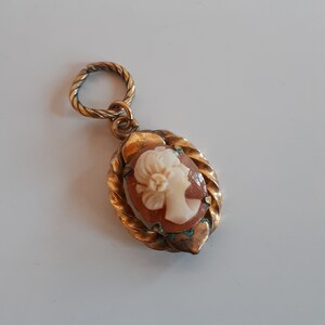May include: A gold-toned cameo pendant featuring a white cameo of a woman's profile with a flower in her hair. The pendant is attached to a twisted gold chain.