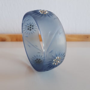 Puede incluir: Un brazalete azul mate con un diseño floral y detalles de diamantes de imitación transparentes.