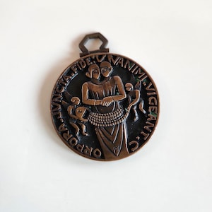 May include: A bronze-colored round pendant with a detailed relief design. The central image depicts two figures with a shared head, surrounded by smaller figures and text. The pendant has a loop at the top for hanging.