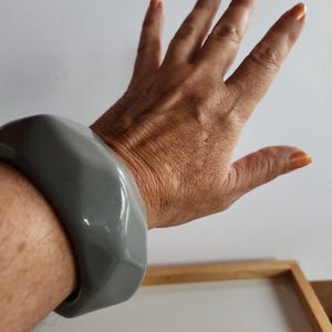 May include: A grey, chunky bracelet with a geometric design. The bracelet is made of a smooth, glossy material and has a rounded, rectangular shape. The bracelet is worn on a wrist.
