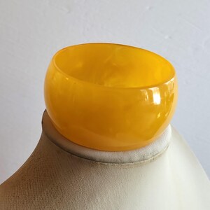 May include: A bright yellow bangle bracelet with a smooth, rounded design. The bracelet has a glossy finish and is made of a solid, translucent material. The colour is a vibrant, sunny yellow. The bangle is displayed on a neutral-coloured surface.