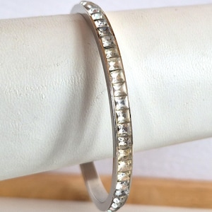 May include: A silver-toned bangle bracelet with a row of square, clear crystals. The crystals are set closely together, creating a continuous sparkle. The bracelet is displayed on a white surface.