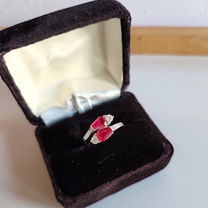 May include: A silver ring with two red rectangular gemstones and two smaller white gemstones, displayed in an open, dark velvet jewellery box. The ring has a modern, elegant wrap-around design.