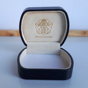 May include: Open black jewelry box with a cream-colored interior. The inside lid features a gold emblem and the text "le Parchemin". The box is empty and ready for a ring or other small jewelry item.