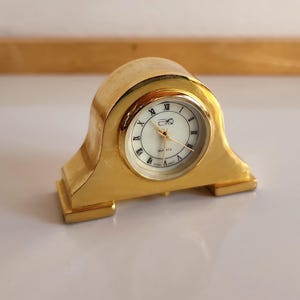 May include: A gold-toned mantel clock with Roman numeral hour markers and a white face. The clock has a decorative base.