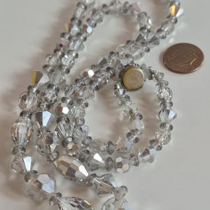 K&ouml;nnte beinhalten: Eine Halskette aus klaren, facettierten Kristallperlen mit abwechselnd runden und l&auml;nglichen Formen und einem irisierenden Finish. Die Kette hat einen runden Metallverschluss und liegt neben einer kupferfarbenen Ein-Cent-M&uuml;nze zum Gr&ouml;&szlig;envergleich.