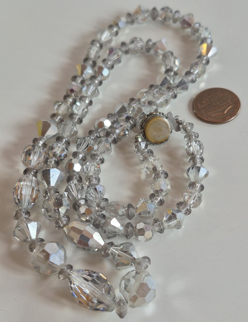 K&ouml;nnte beinhalten: Eine Halskette aus klaren, facettierten Kristallperlen mit abwechselnd runden und l&auml;nglichen Formen und einem irisierenden Finish. Die Kette hat einen runden Metallverschluss und liegt neben einer kupferfarbenen Ein-Cent-M&uuml;nze zum Gr&ouml;&szlig;envergleich.
