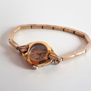 Könnte beinhalten: Eine goldfarbene Armbanduhr mit einem runden Zifferblatt und einem zarten, blattartigen Design am Armband.