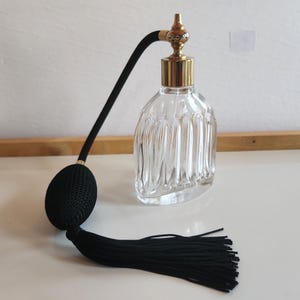 May include: A clear glass perfume bottle with a gold-coloured top and a black bulb atomizer. The bottle has a ribbed design and a black tassel. The atomizer has a black rubber tube and a black bulb.