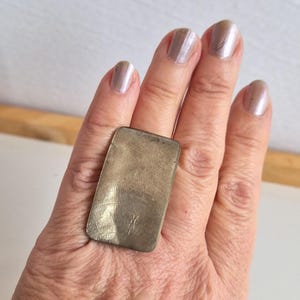 May include: A silver rectangular ring with a textured surface. The ring is worn on a hand with polished nails. The ring's design is simple and modern, making it a versatile accessory.