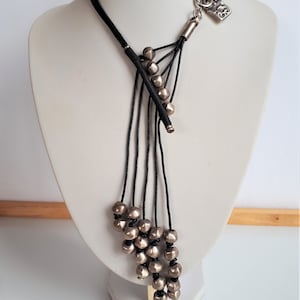 Pode incluir: Um colar longo de cord&atilde;o preto com um fecho de cadeado prateado e contas prateadas. As contas est&atilde;o agrupadas na parte inferior do colar, criando um efeito em cascata.