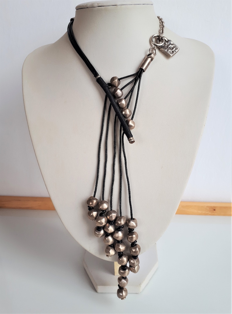 Pode incluir: Um colar longo de cord&atilde;o preto com um fecho de cadeado prateado e contas prateadas. As contas est&atilde;o agrupadas na parte inferior do colar, criando um efeito em cascata.