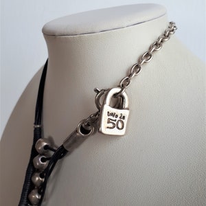 Pode incluir: Um colar de couro preto com contas prateadas e um pingente de cadeado prateado. O cadeado tem o texto "Uno de 50" gravado.