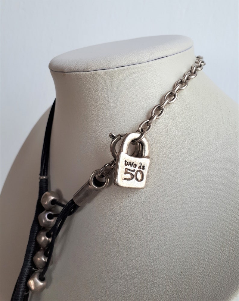 Pode incluir: Um colar de couro preto com contas prateadas e um pingente de cadeado prateado. O cadeado tem o texto "Uno de 50" gravado.