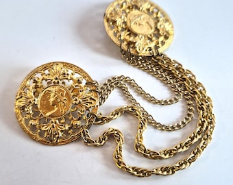 Könnte beinhalten: Vintage goldfarbene Brosche mit zwei runden Filigranmedaillons, die durch Ketten verbunden sind. Jedes Medaillon zeigt ein Profilporträt in einem aufwendigen, durchbrochenen Design. Ein klassisches Accessoire.