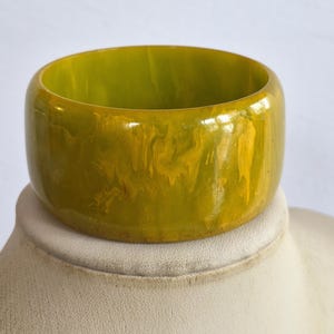 May include: A wide, circular bangle bracelet featuring a polished, smooth surface with a marbled pattern of olive green and bright yellow swirls. The thick, solid band has a glossy finish and a consistent, rounded shape throughout its entire circumference.