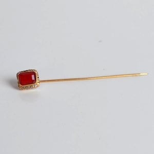 May include: A gold stick pin featuring a rectangular, red gemstone. The gemstone is set in a gold frame with small decorative details. The pin is presented on a white surface.