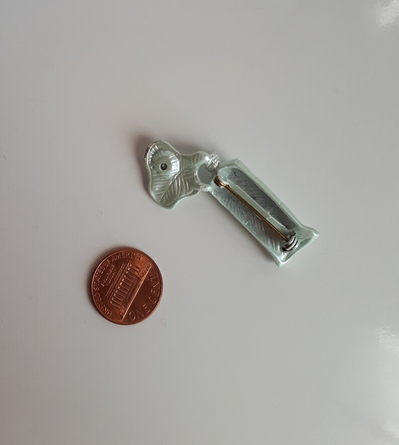 May include: A light green metal pin with a leaf design and a pin back. The pin is about the size of a US penny.