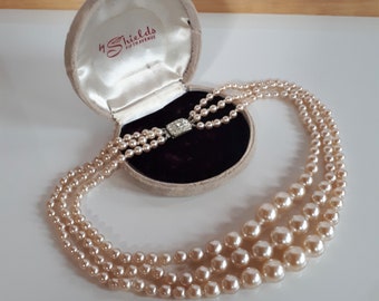 May include: A three-strand pearl necklace with a rhinestone clasp, displayed outside of a vintage jewelry box. The box is beige with the text "4 Shields Fifth Avenue" printed on the inside.