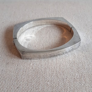 May include: A silver bangle bracelet with a square shape and a hinged closure.